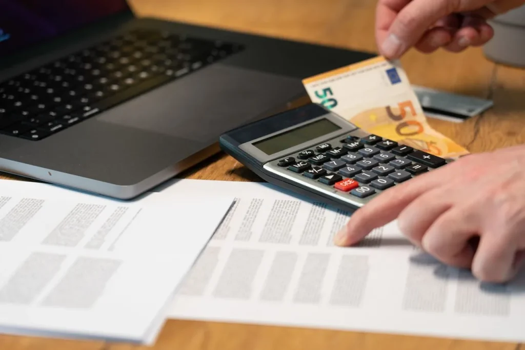 Ręce liczące na kalkulatorze przy banknotach euro i laptopie - przeliczanie prowizji Airbnb 15,5%